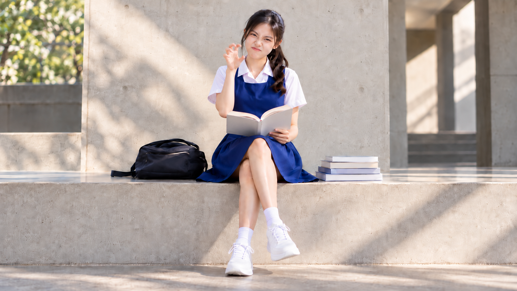 A student in a blue-and-white uniform sits with an open book on a sunlit stone bench, with a bag and a small stack of books beside her.