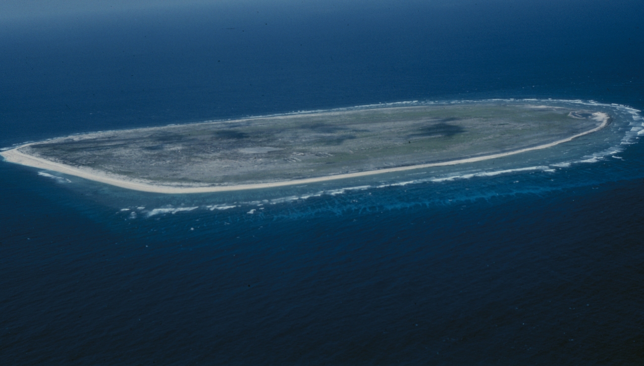 Aerial view of Baker Island’s low, pale shoreline and reef edge surrounded by deep blue Pacific water.