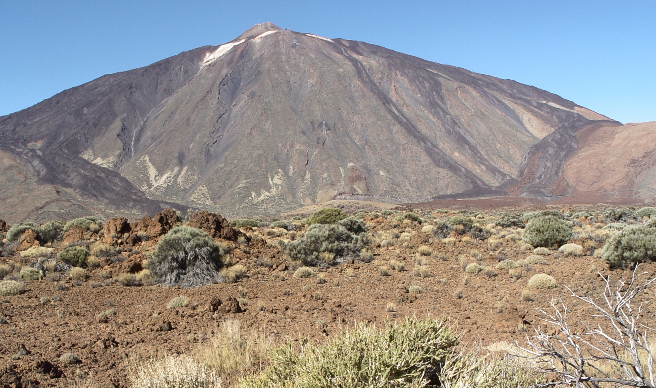 The volcanic cone of Mount Teide rises above brown lava rock and hardy mountain shrubs under a clear sky in Tenerife, Canary Islands.