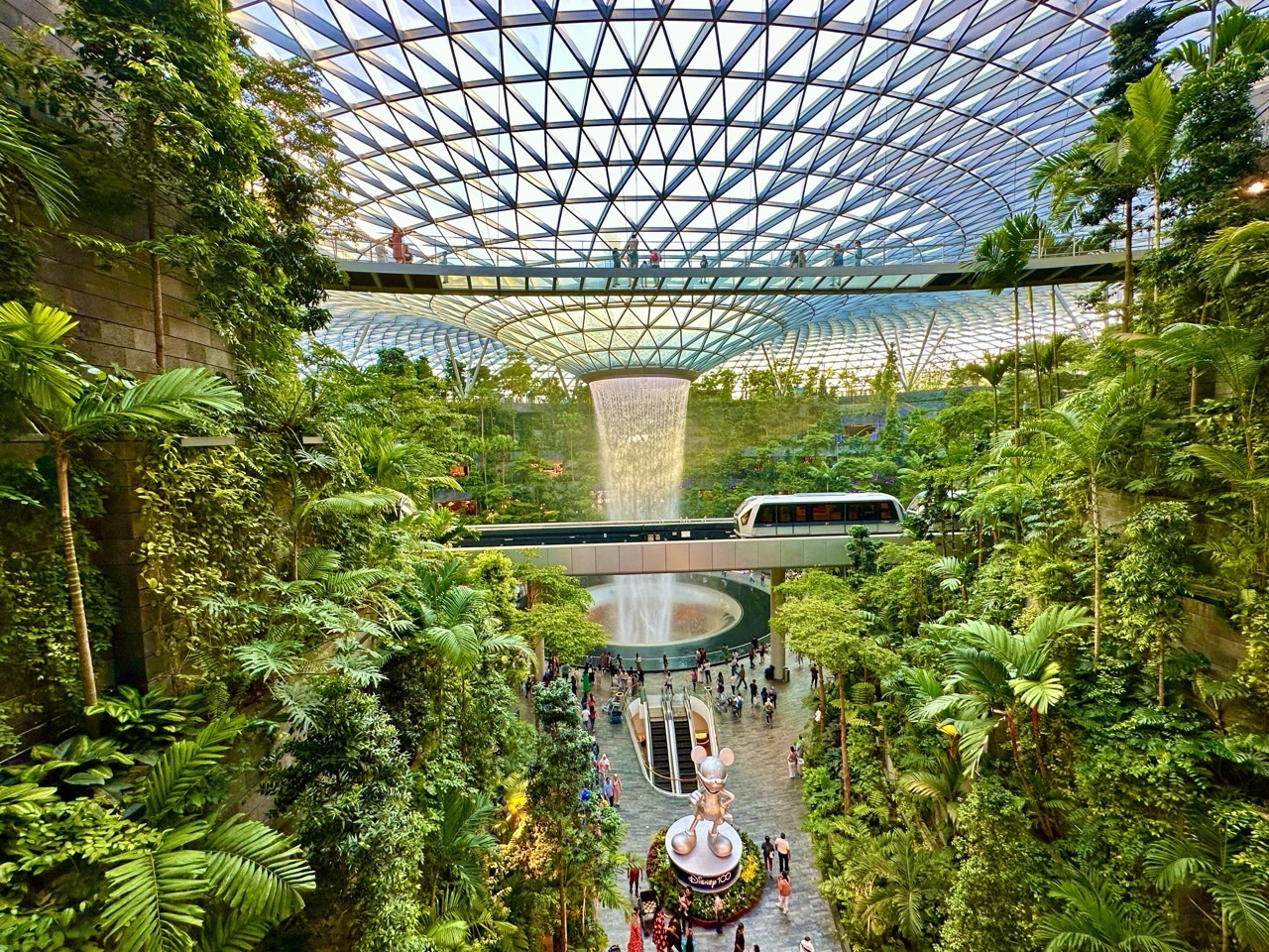 Jewel Changi Airport interior with roof structure, foliage, waterfall, glass-floor bridge and Skytrain passing through the dome.