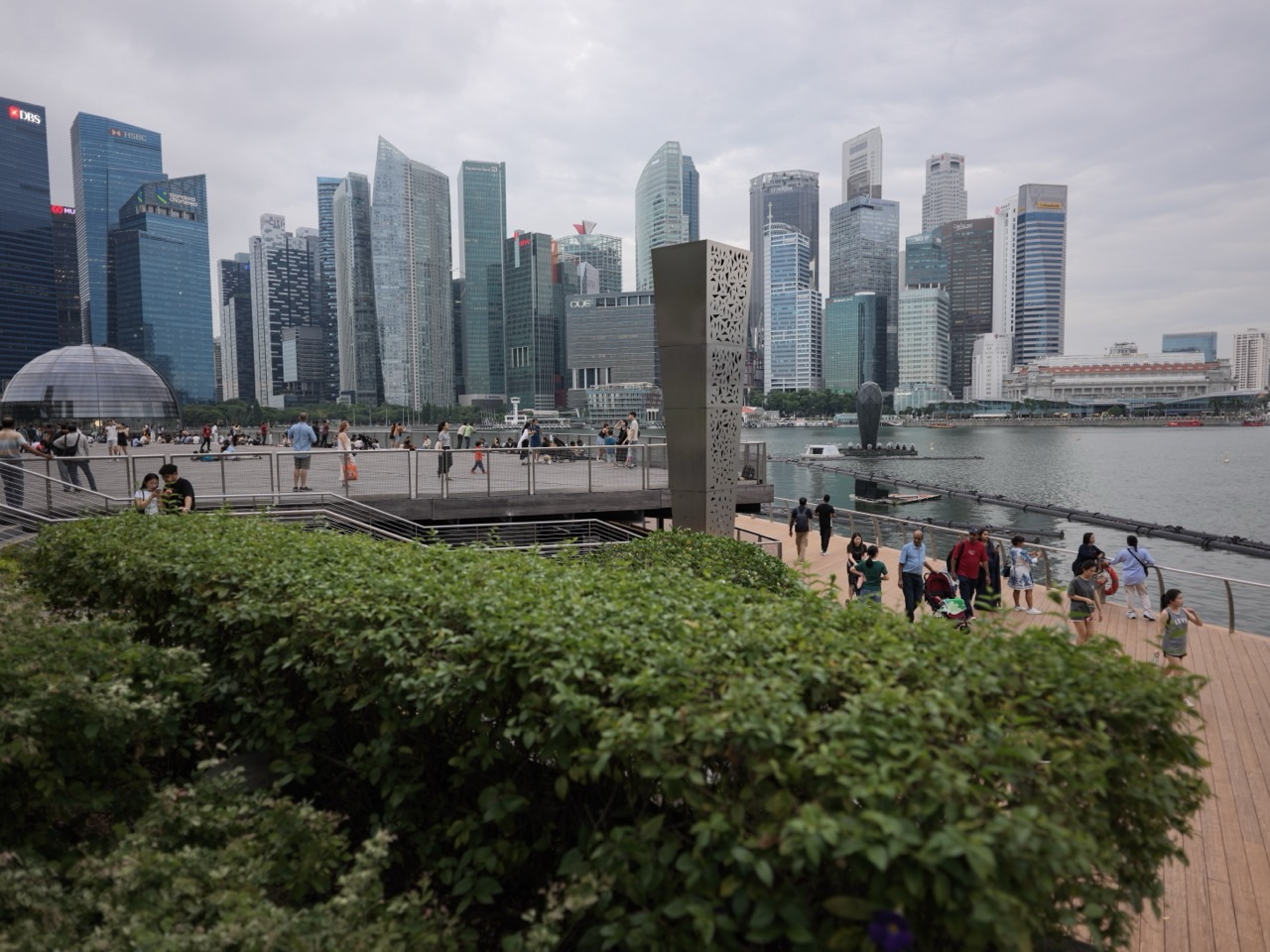 Tourists and shoppers walking along the waterfront promenade outside The Shoppes at Marina Bay Sands with the financial district skyline behind