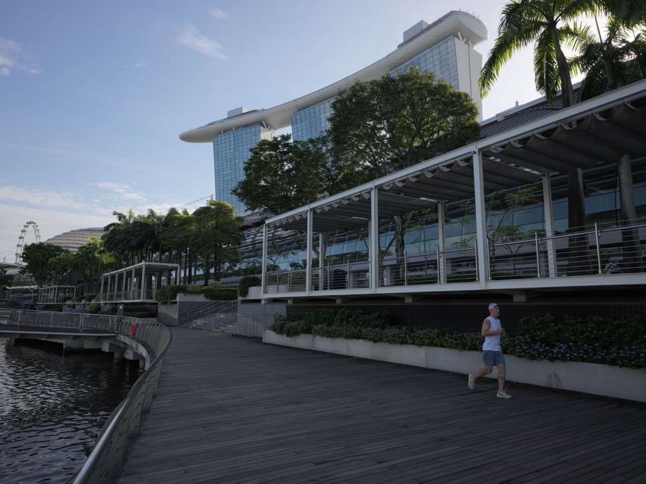 Runner on the waterfront promenade outside The Shoppes at Marina Bay Sands, with roof architecture, hotel towers and the SkyPark behind
