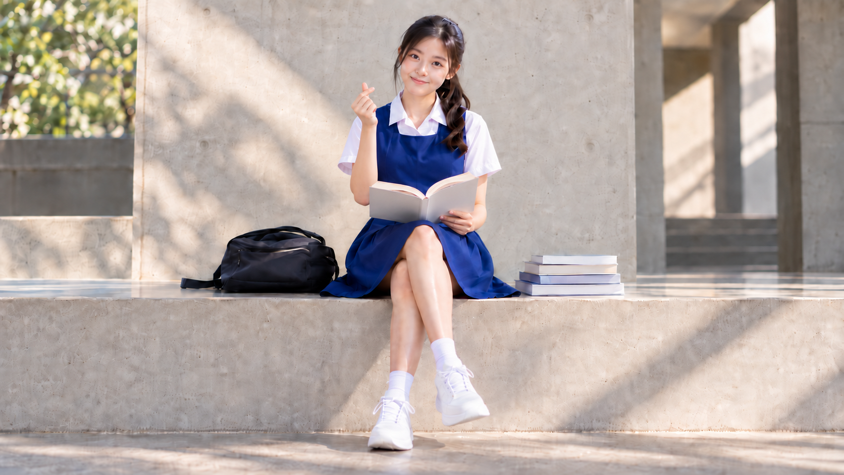 Student in a blue pinafore reads an open book on a sunlit stone bench beside a black bag and stacked books.