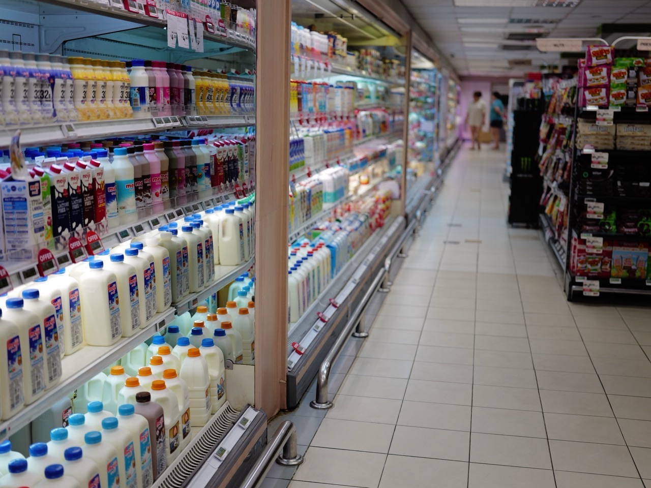 Shoppers in a chilled supermarket dairy aisle with milk products on display.