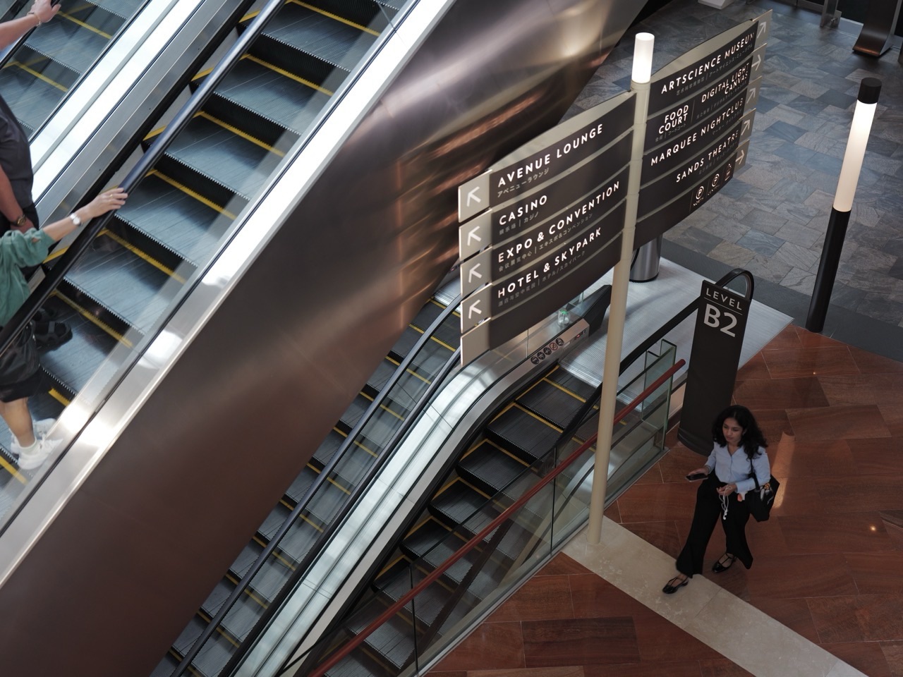 Overhead view of a visitor walking beside an escalator and direction signs at Marina Bay Sands.
