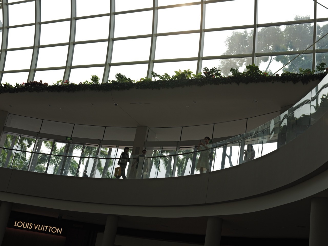 Silhouetted shoppers beneath an interior roof at Marina Bay Sands, with trees visible outside.
