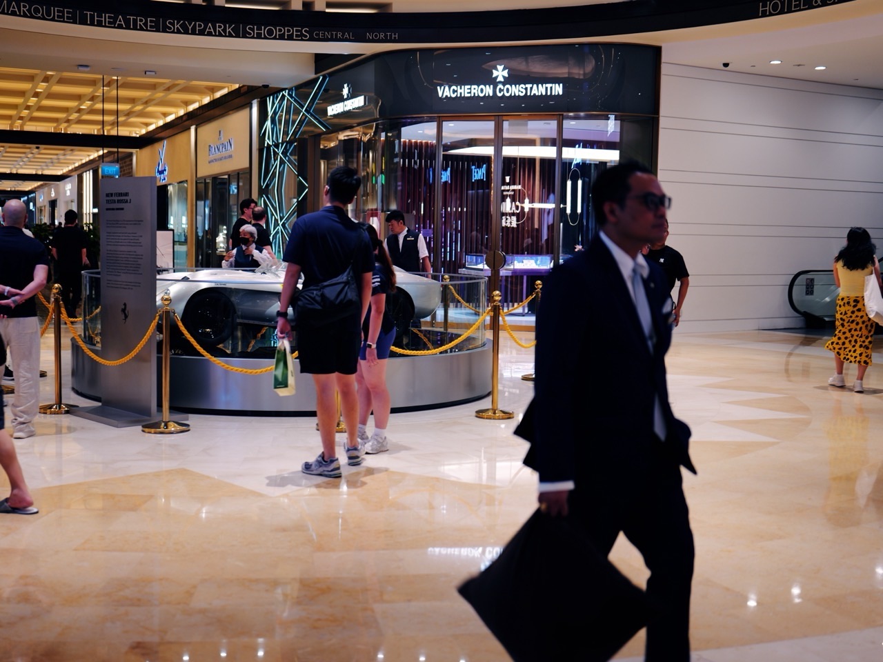 A suited visitor among shoppers in a Marina Bay Sands foyer, with a car display in the background.