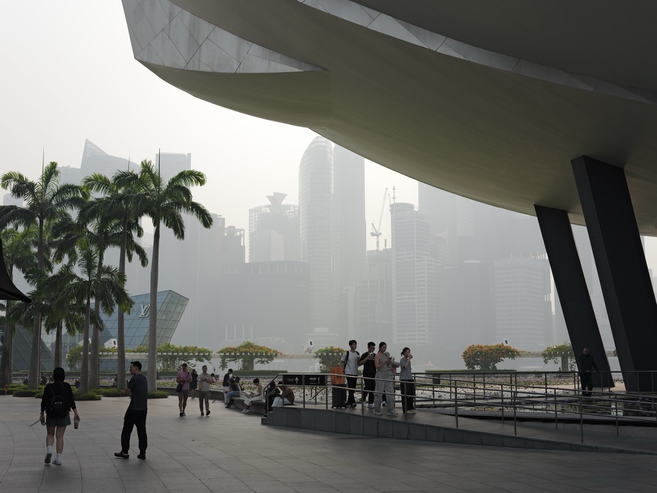 Pedestrians between ArtScience Museum on the right and a hazy Singapore skyline on the left.