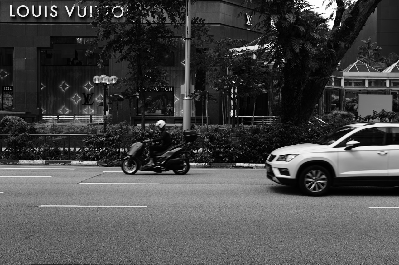 Ngee Ann City luxury storefronts with a car and motorcyclist on Orchard Road in Singapore.