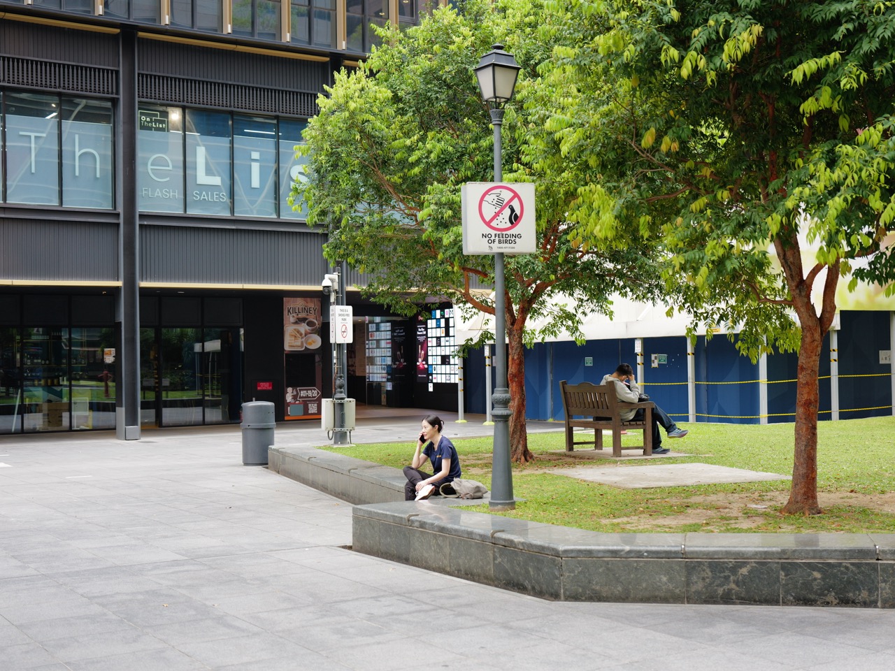 Woman using her phone near a park sign and security camera, with another man nearby.