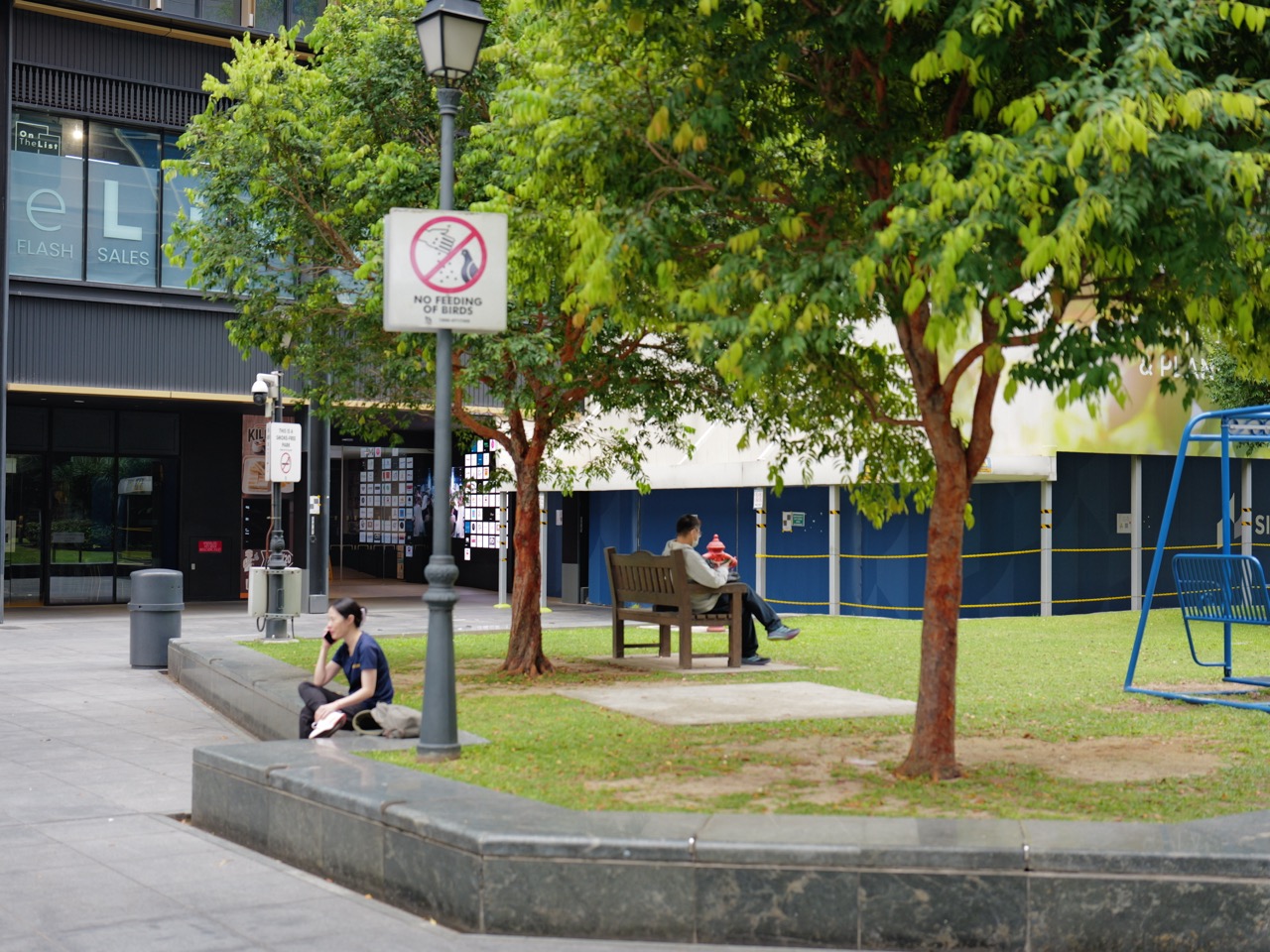 Woman using a mobile phone near another park visitor, bird-feeding signage and a surveillance camera.