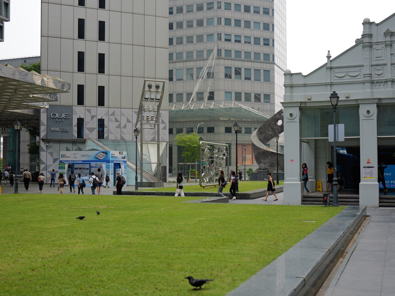 Commuters walking out of Raffles Place MRT with OUE and UOB office buildings and public art behind.