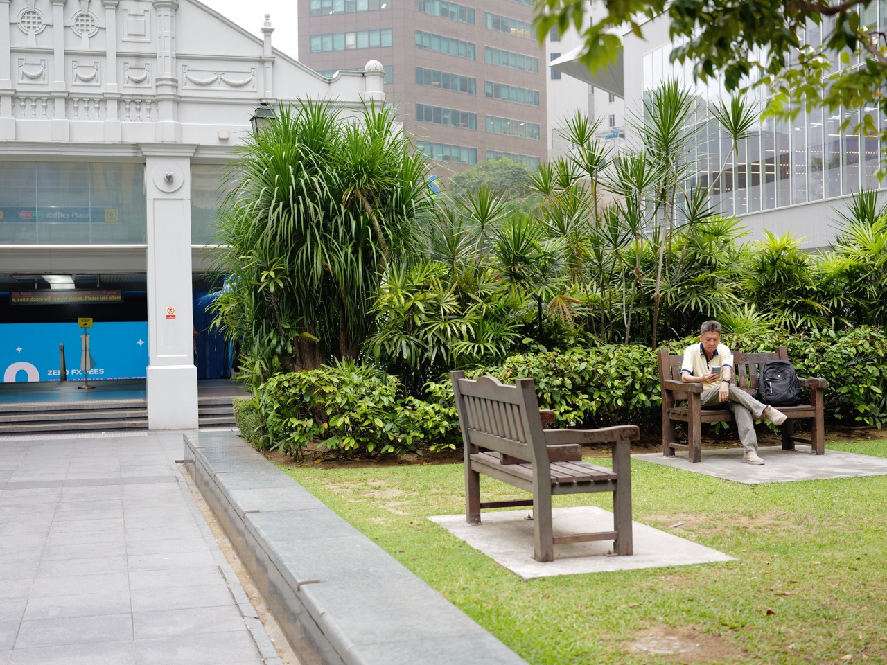 Man sitting in a park and looking at his phone near the Raffles Place MRT exit.
