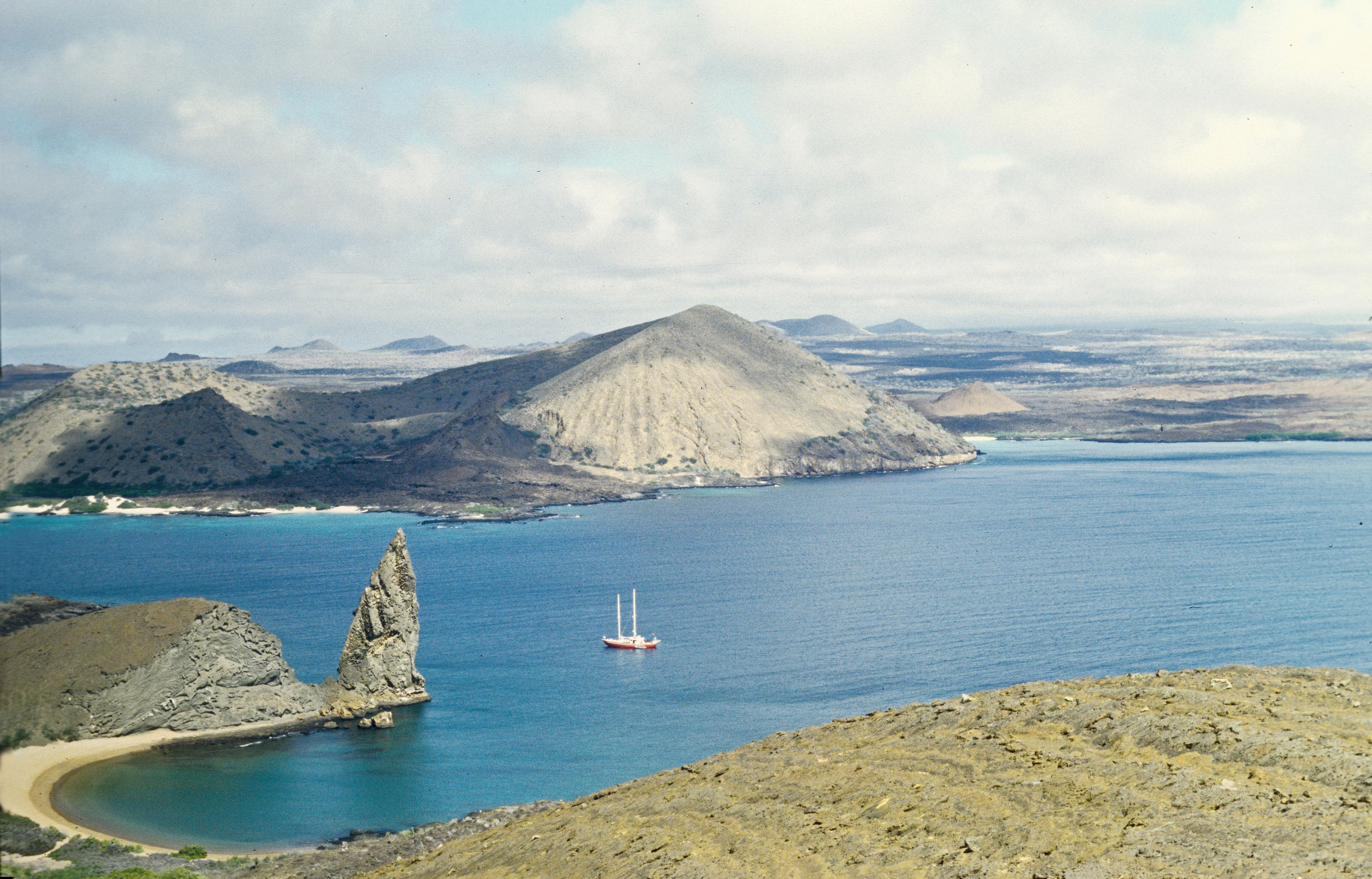 View over the volcanic coast of Bartolomé Island with the pointed Pinnacle Rock and a boat offshore in the Galápagos Islands, photographed in 2002.