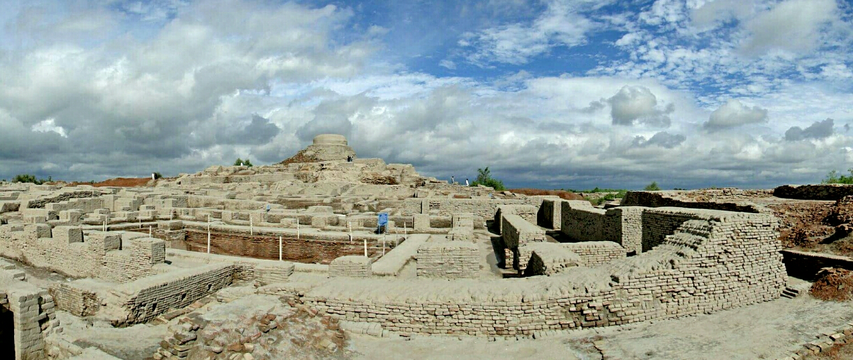 Wide photograph of excavated brick street blocks and buildings at Mohenjo-daro in Sindh, Pakistan, an ancient Indus city.