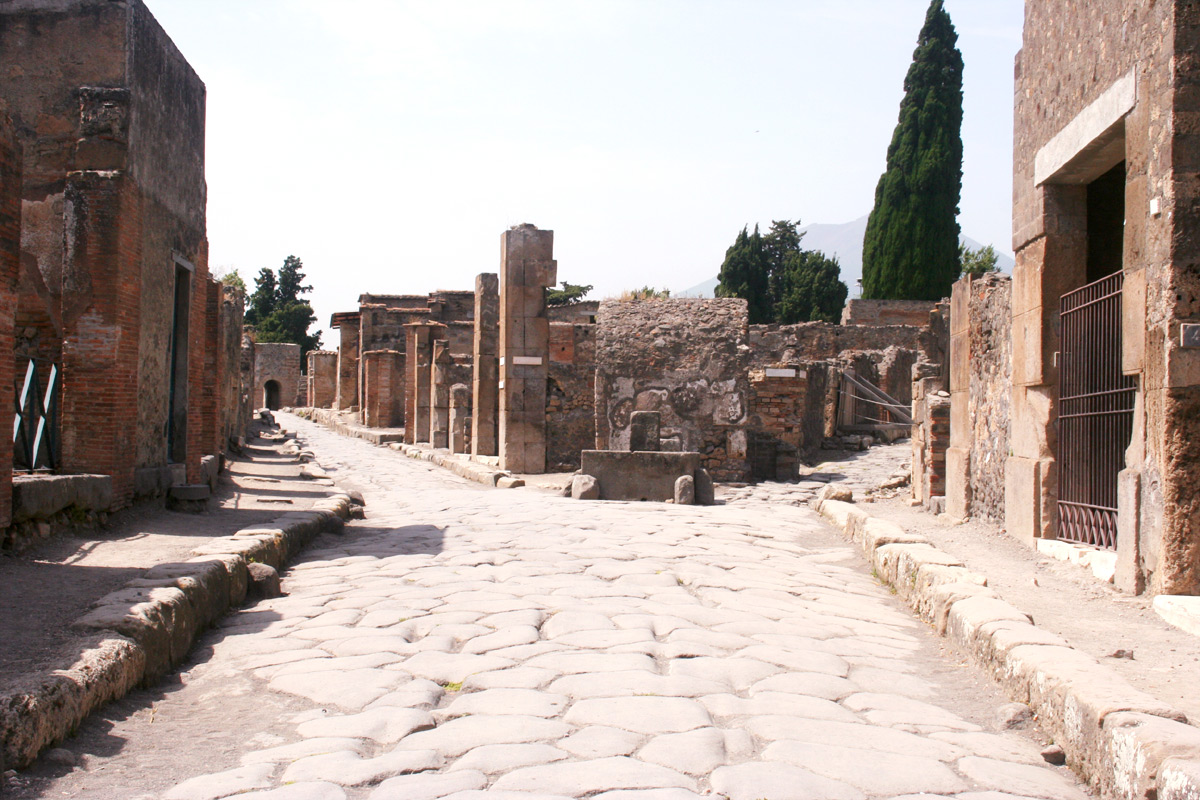 A surviving stone-paved street at the archaeological city of Pompeii in Italy, with ancient buildings and ruins on both sides.